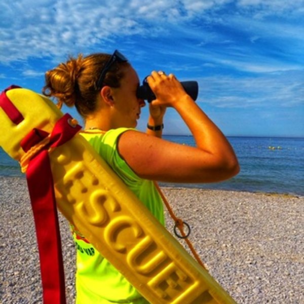 Lily-Marie sauveteuse sur une plage de galets, une bouée tube sur l'épaule, surveillant la mer aux jumelles